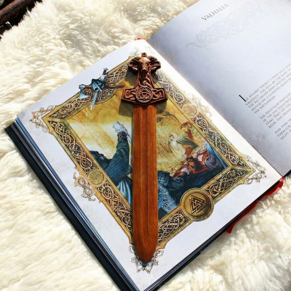Hand-carved wooden Viking sword bookmark with an ornate ram-head hilt and Celtic knots. Pictured on an open mythology book, this artisan-crafted piece is an ideal luxury gift for Norse history lovers and collectors of fine woodwork.